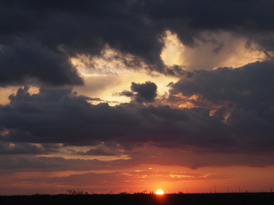 [Two-thirds of the yellow-orange orb sits above the flat, dark horizon. The clouds in the immediate background of are shades of orange. The clouds above those are dark blue. There is a section in the top which is open to the west and thus those clouds are still white as they are lit by the sun in the distance.]